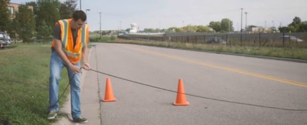 Field worker in orange safety vest laying down a pneumatic tube for data collection. Two orange traffic cones are in the background